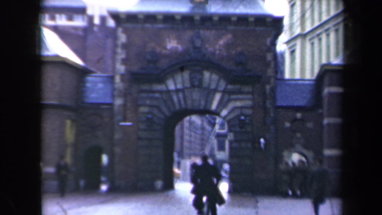 1940s Netherlands home-movie frame showing a large stone archway, pedestrians, and a cyclist moving through an urban street.