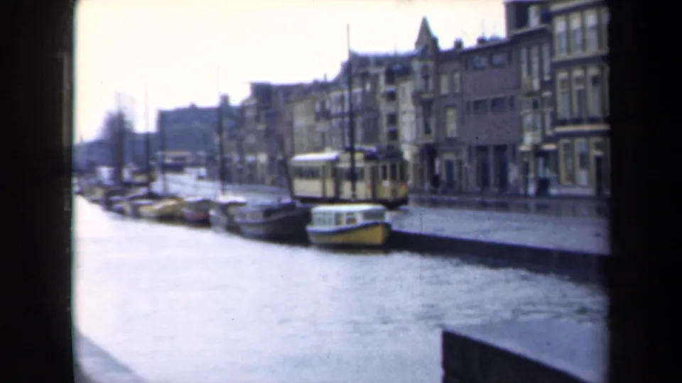 Canal-side Dutch street with small boats, waterfront buildings, and a tram visible along the water's edge.