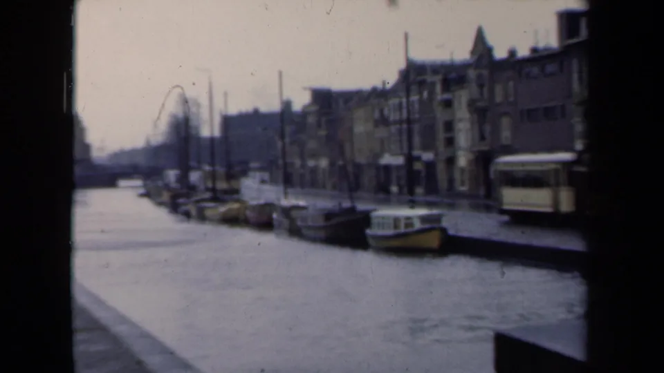 1946 Netherlands canal view from a handheld 8mm camera, with moored boats, older buildings, and a tram at the street edge.