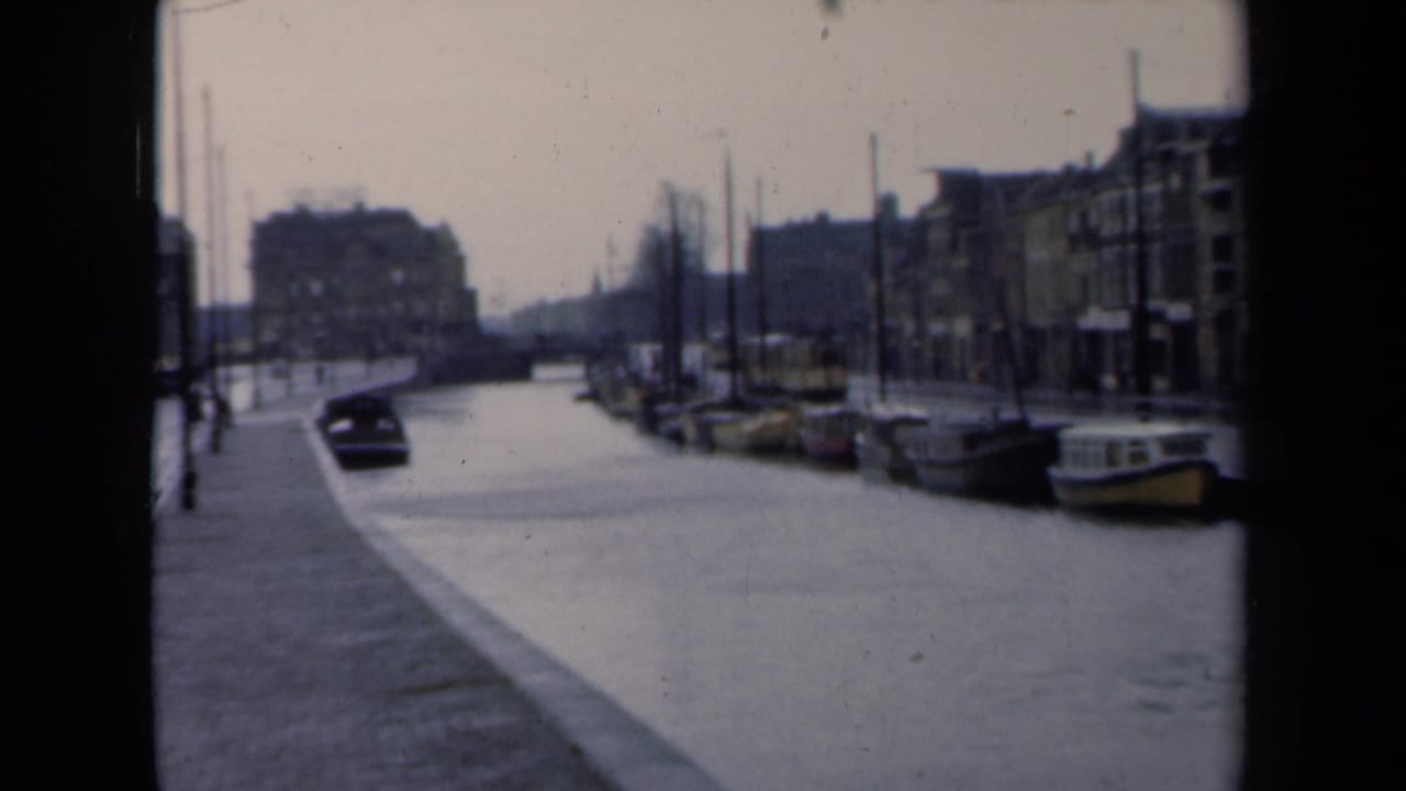 1946 Netherlands canal street with wet roadway, parked boats, a dark car, lampposts, and soft-focus urban buildings.