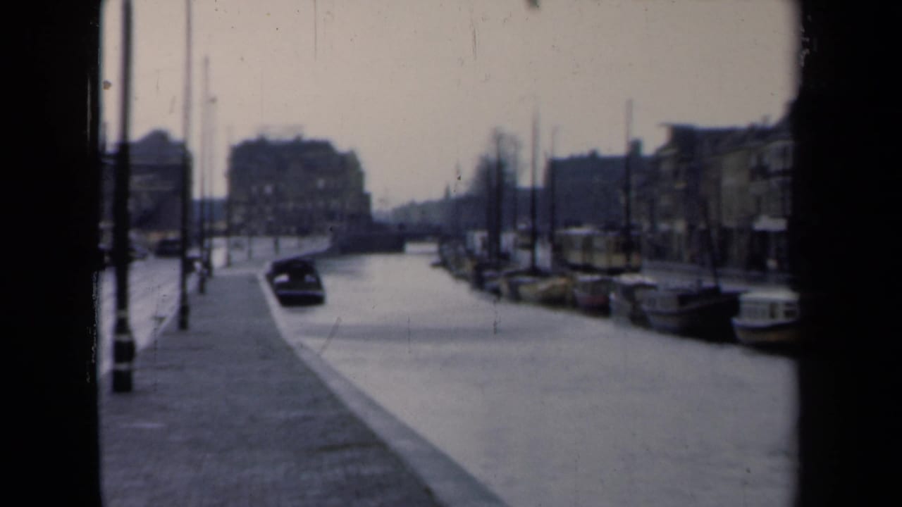 Canal view with a dark car, moored boats, wet street, lampposts, and buildings along the right side.