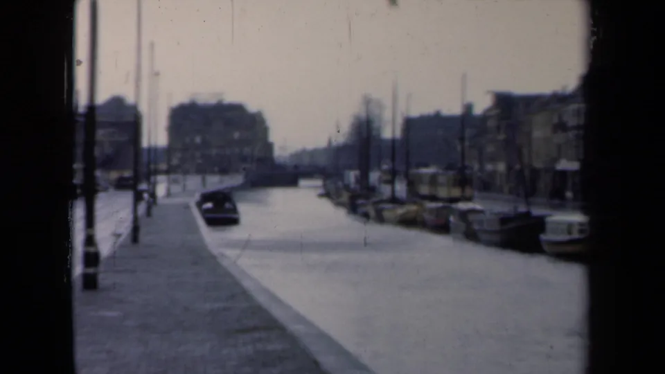 Soft 8mm view of a Netherlands canal street in 1946, showing a dark car, moored boats, wet pavement, and waterfront buildings.