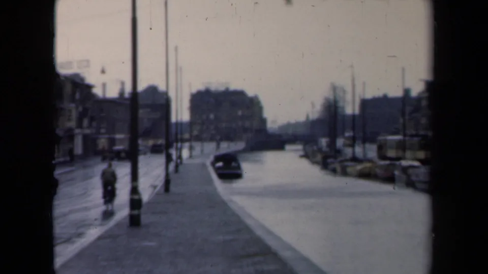 1946 Netherlands canal-side street with a small visible person, dark car, wet paving, lampposts, boats, and blurred urban architecture.