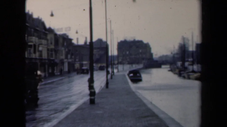Soft 1946 Netherlands street frame showing canal-side traffic, moored boats, wet paving, lampposts, storefront-like facades, and distant people.