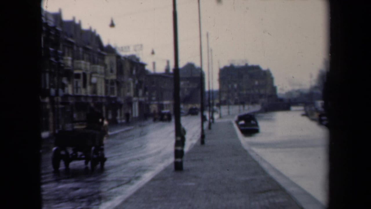1940s Netherlands home-movie frame showing a horse-drawn carriage on a wet urban street with lampposts and multi-story buildings.