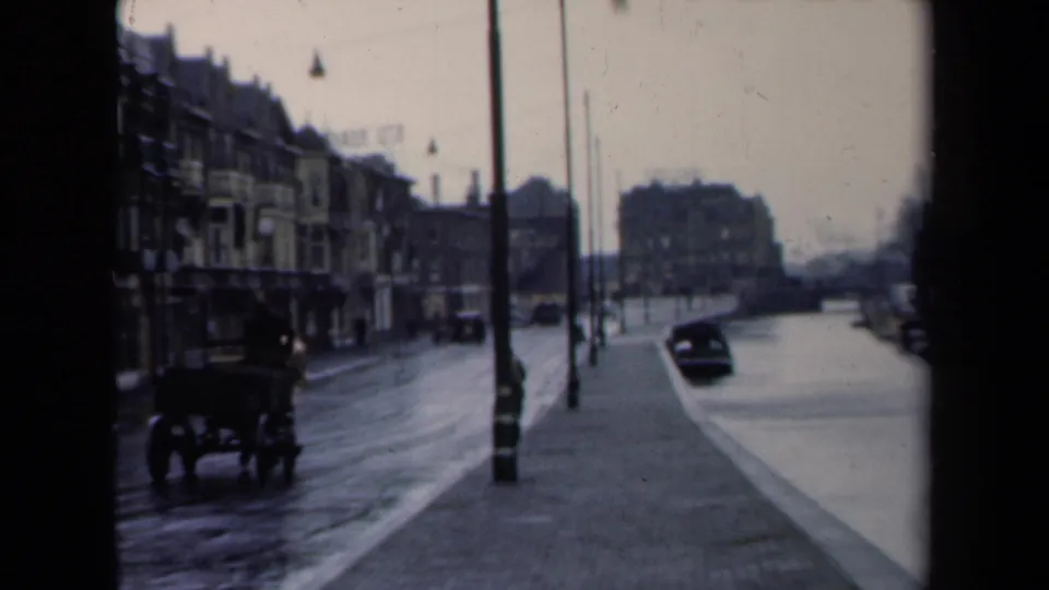 1940s Netherlands home-movie frame showing a horse-drawn carriage on a wet urban street with lampposts and multi-story buildings.