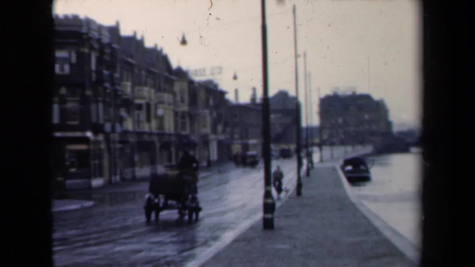 1940s Netherlands street view with a horse-drawn carriage, wet pavement, motorcars, lampposts and a small distant figure.