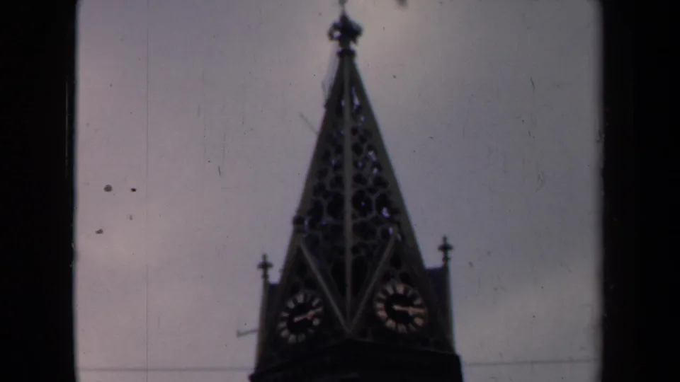 Archival 8mm view of a pointed clock tower with two clock faces, muted exposure and visible physical-film texture.