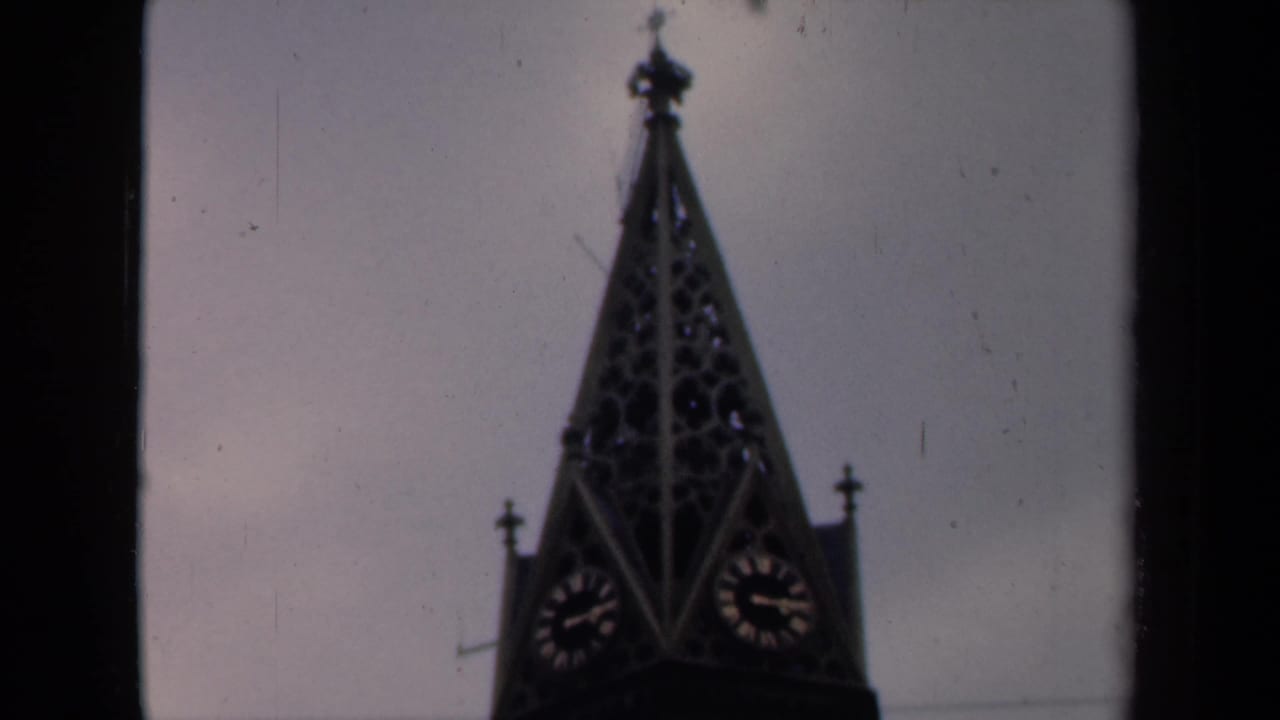 1946 Netherlands home-movie frame showing a dark church spire with clock faces under an overcast sky.