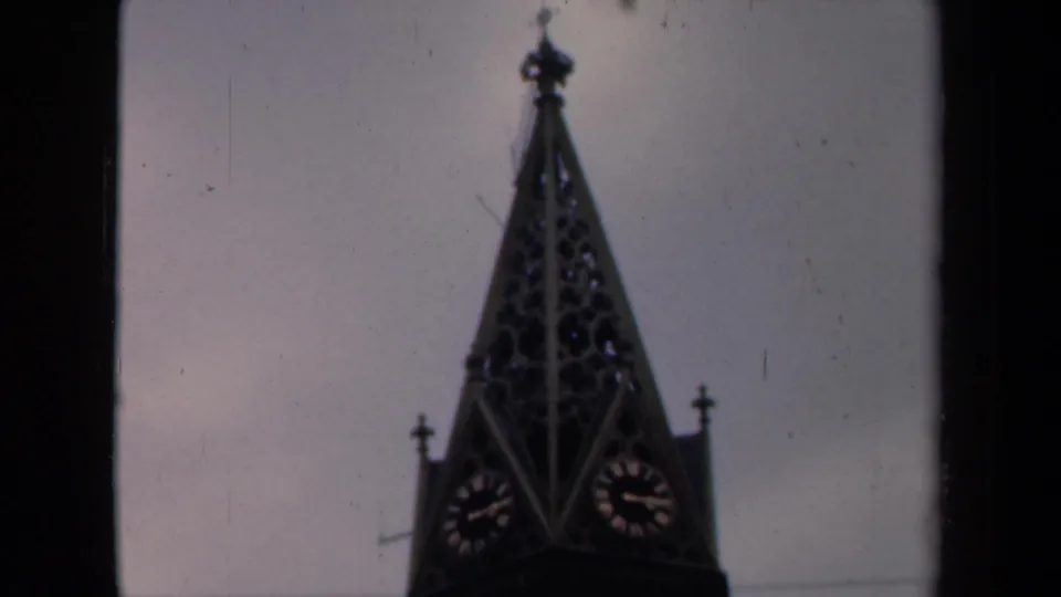 1946 Netherlands home-movie frame showing a dark church spire with clock faces under an overcast sky.