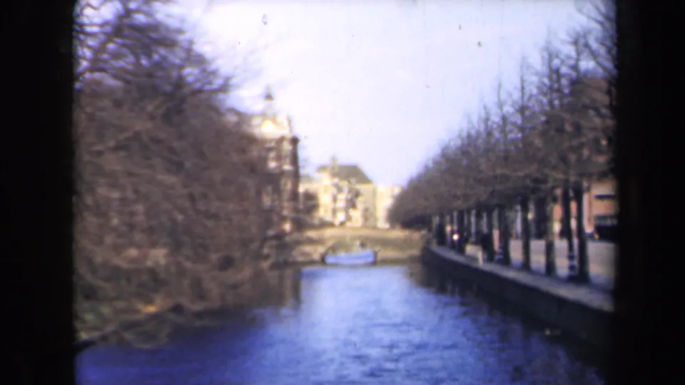 1946 Netherlands canal view with leafless trees, distant masonry buildings, and a small boat on the water.