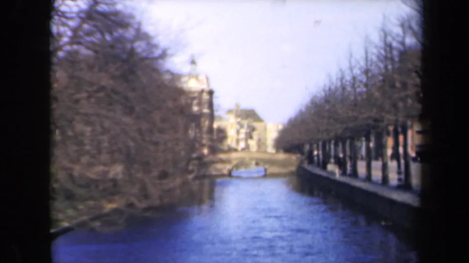 1946 Netherlands home-movie frame with blue-toned canal water, bare trees, and distant waterside buildings.
