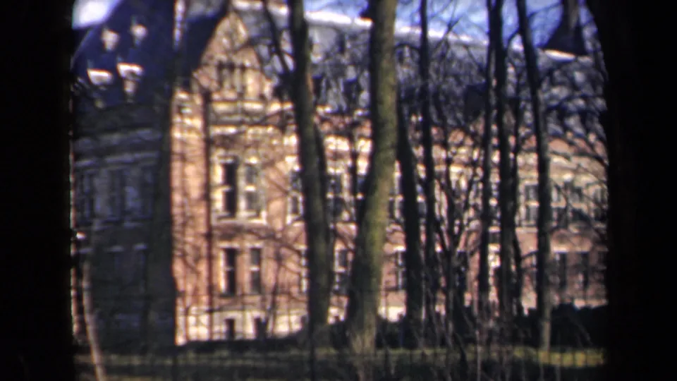 Soft 8mm frame from the Netherlands showing a tall brick building corner behind bare tree trunks.