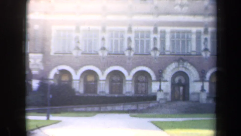 1946 Netherlands 8mm frame of a formal masonry building facade with arched lower openings, rows of windows, lawn, and walkway.