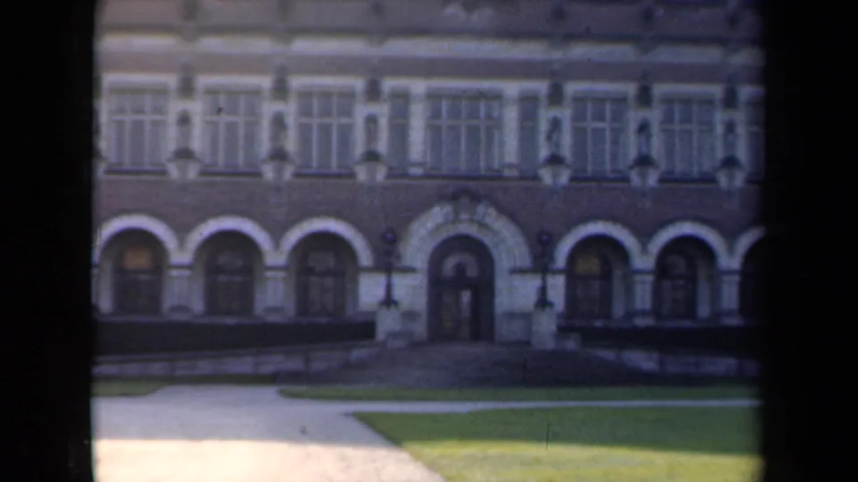 1946 Netherlands 8mm frame of a broad formal facade with a central arched doorway, repeated windows, hedges, and green lawn in front.