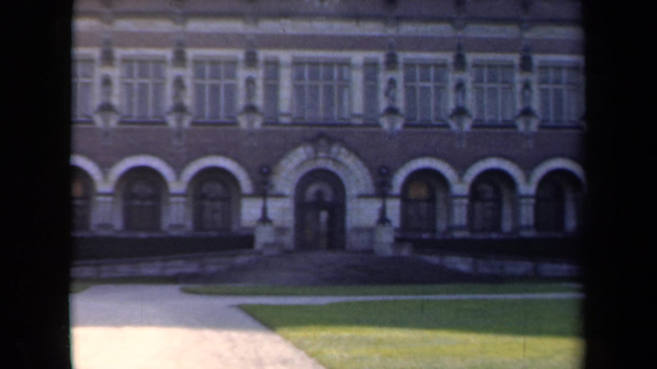 1946 Netherlands 8mm frame showing a formal brick-and-stone building facade with arched entries, tall windows, lawn, and paths.