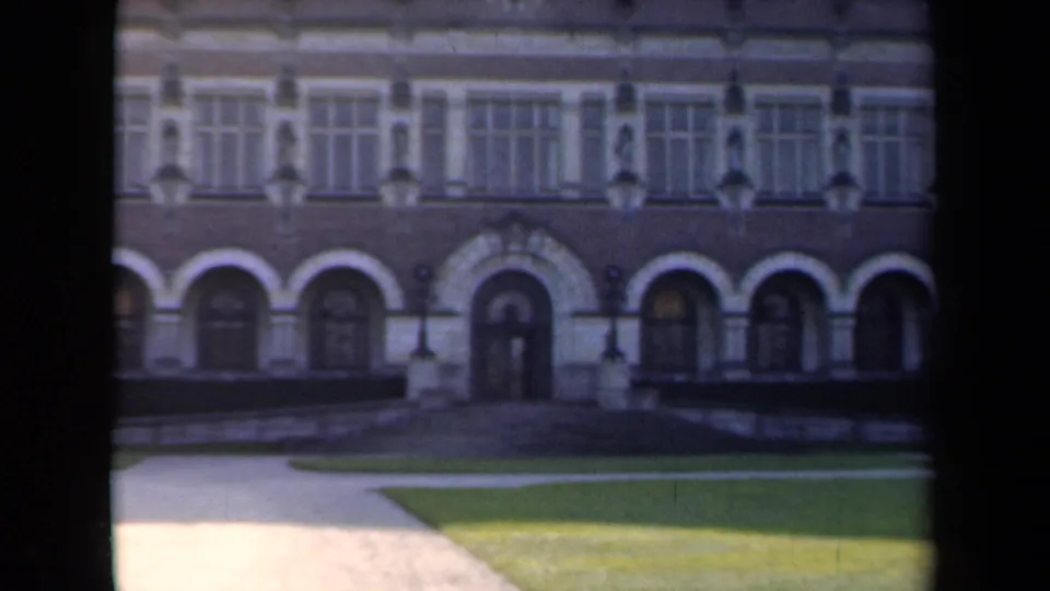 1946 Netherlands 8mm frame showing a formal brick-and-stone building facade with arched entries, tall windows, lawn, and paths.