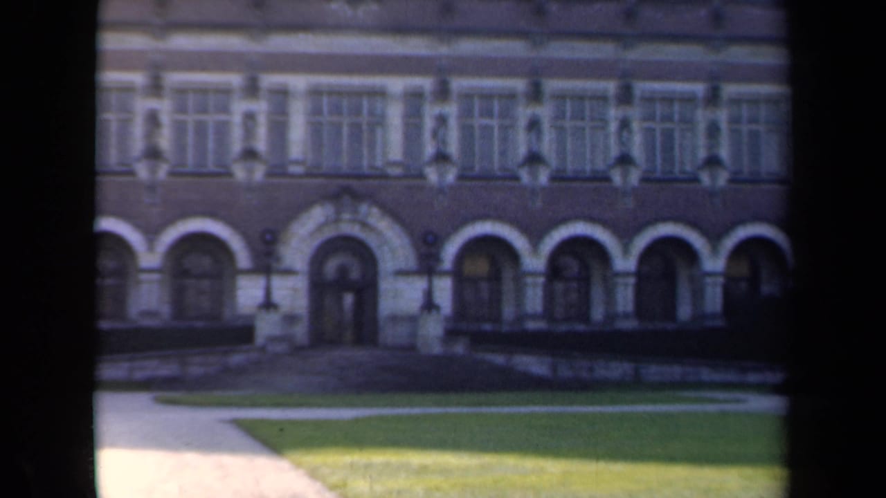 Building front with repeated arches, upper windows, shadowed lawn, and walkway.