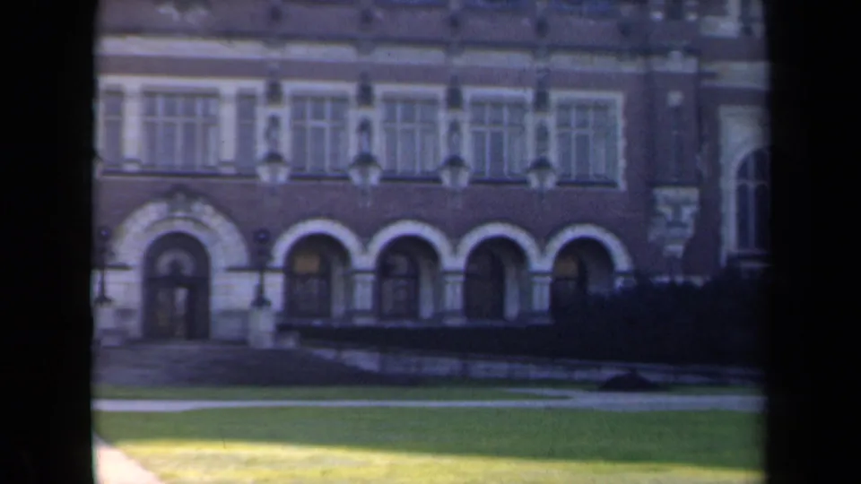 1946 Netherlands 8mm frame showing the building facade with arches, rows of windows, trimmed hedge, sunlit lawn, and a pale path.