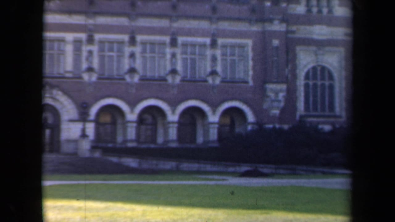 1946 Netherlands home-movie frame showing a formal historic building facade beyond a grassy lawn, with arched openings and soft film grain.