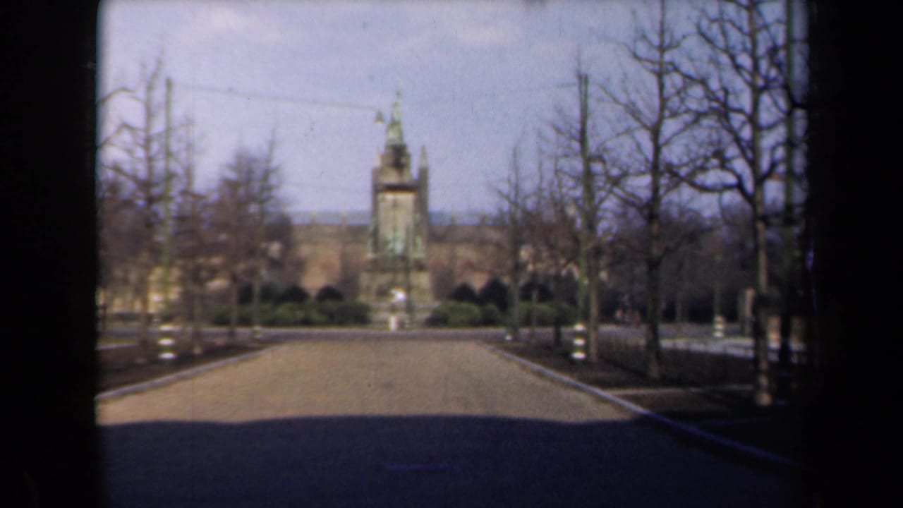 Broad road with bare trees and a tall monument centered beyond landscaped grounds.
