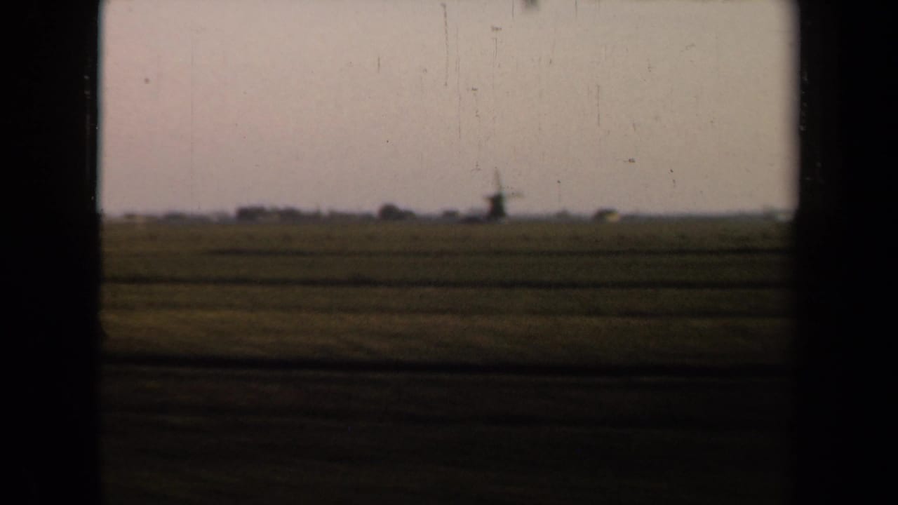 Flat farmland seen through dark train-window framing, with a distant windmill on the horizon.