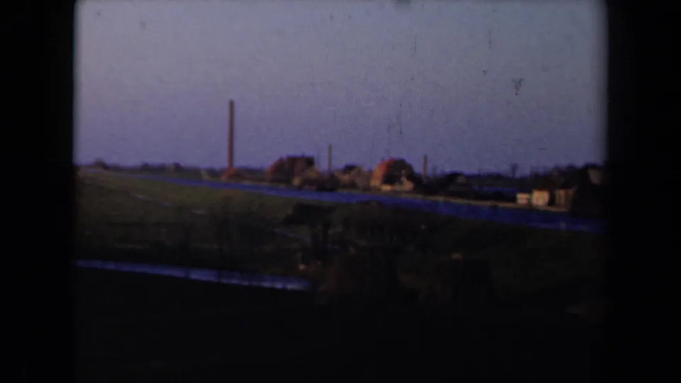 1946 Netherlands 8mm frame showing a rural canal-side settlement, low houses, green fields, and a tall chimney seen from a moving train.