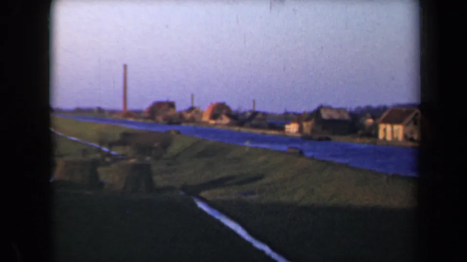 Netherlands 1946 home-movie frame of a rural waterway with grassy banks, small houses, and a tall chimney under a blue sky.