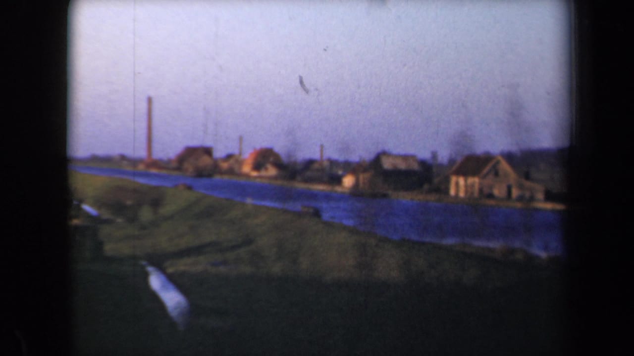 Blue waterway beside grassy banks and low village houses under a pale sky.