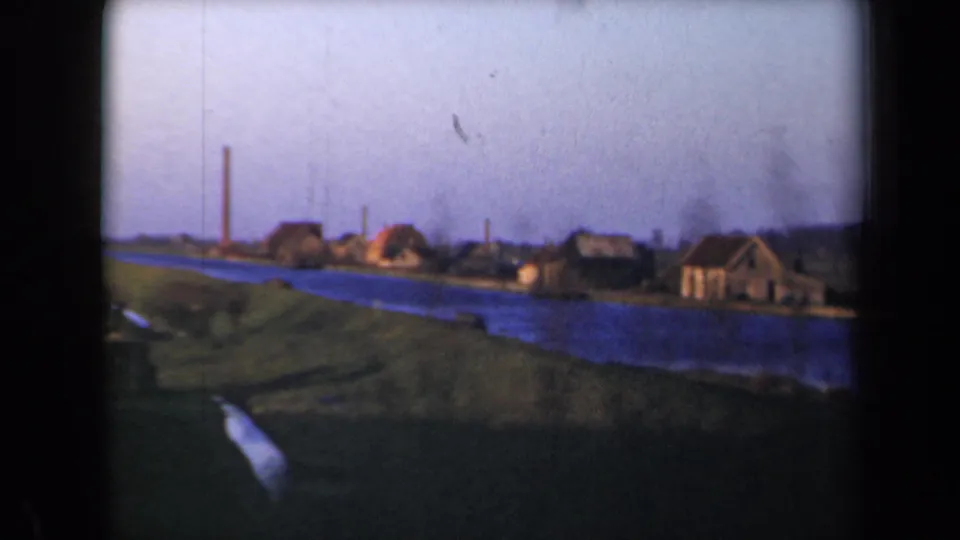 1946 Netherlands home-movie frame showing a blue canal or river, grassy bank, thatched houses, and a distant chimney.
