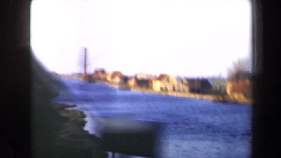 1946 Netherlands frame with a bright sky, blue canal or river, grassy near bank, distant houses, and a tall chimney.