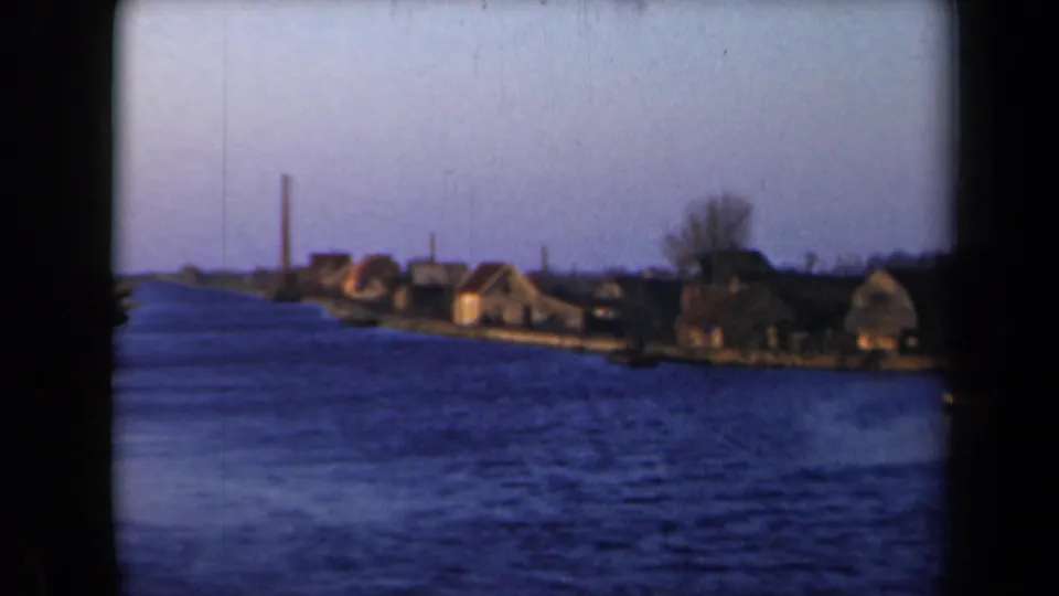 1946 Netherlands home-movie frame showing rippled blue water, thatched homes, bare trees, and a tall chimney beyond the bank.