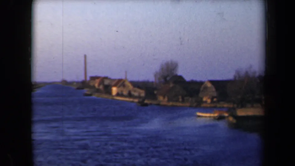 1946 Netherlands frame showing a blue waterway, low waterside houses, bare trees, a tall chimney, and a small boat at right.
