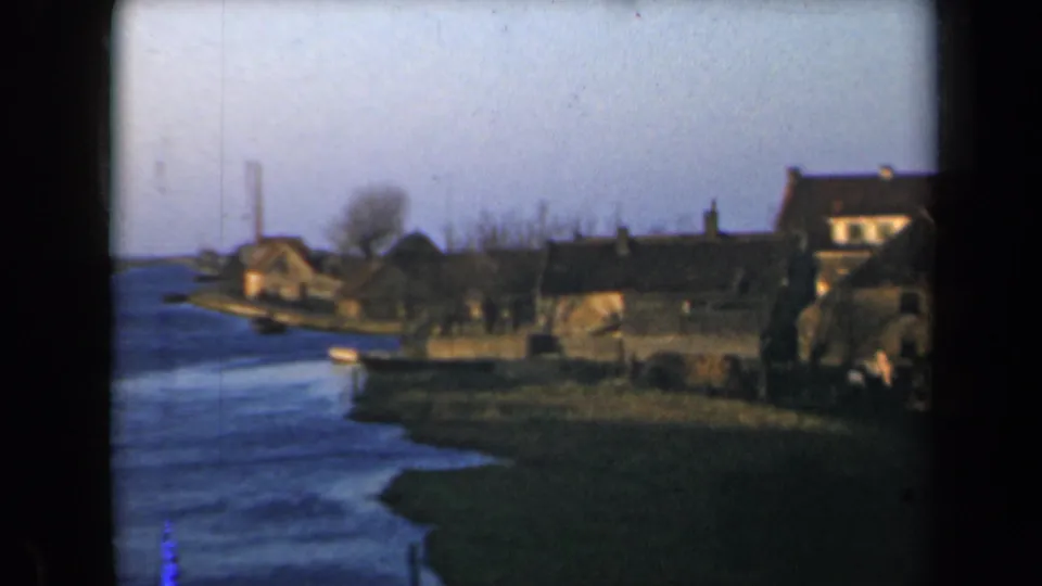 1946 Netherlands 8mm frame with brown riverside houses, a grassy bank, bare trees, and water curving along the village edge.