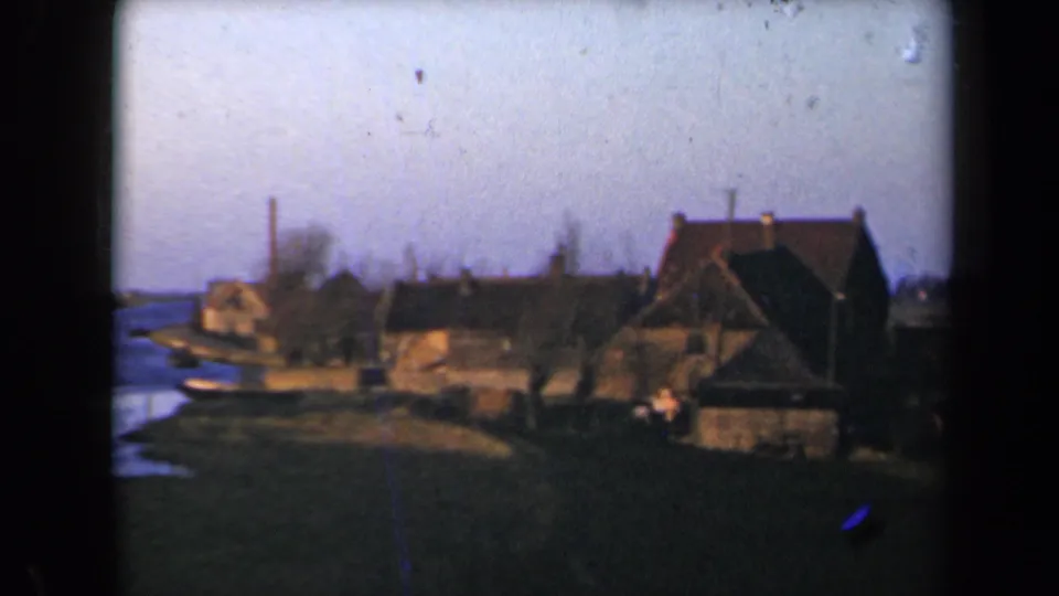 1946 Netherlands 8mm frame showing a waterside village with thatched roofs, bare trees, grassy foreground, and water at left.