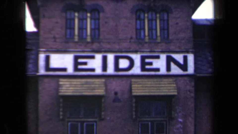 1946 Netherlands frame with soft 8mm texture showing a white LEIDEN sign, brick facade, arched upper windows, and striped awnings.
