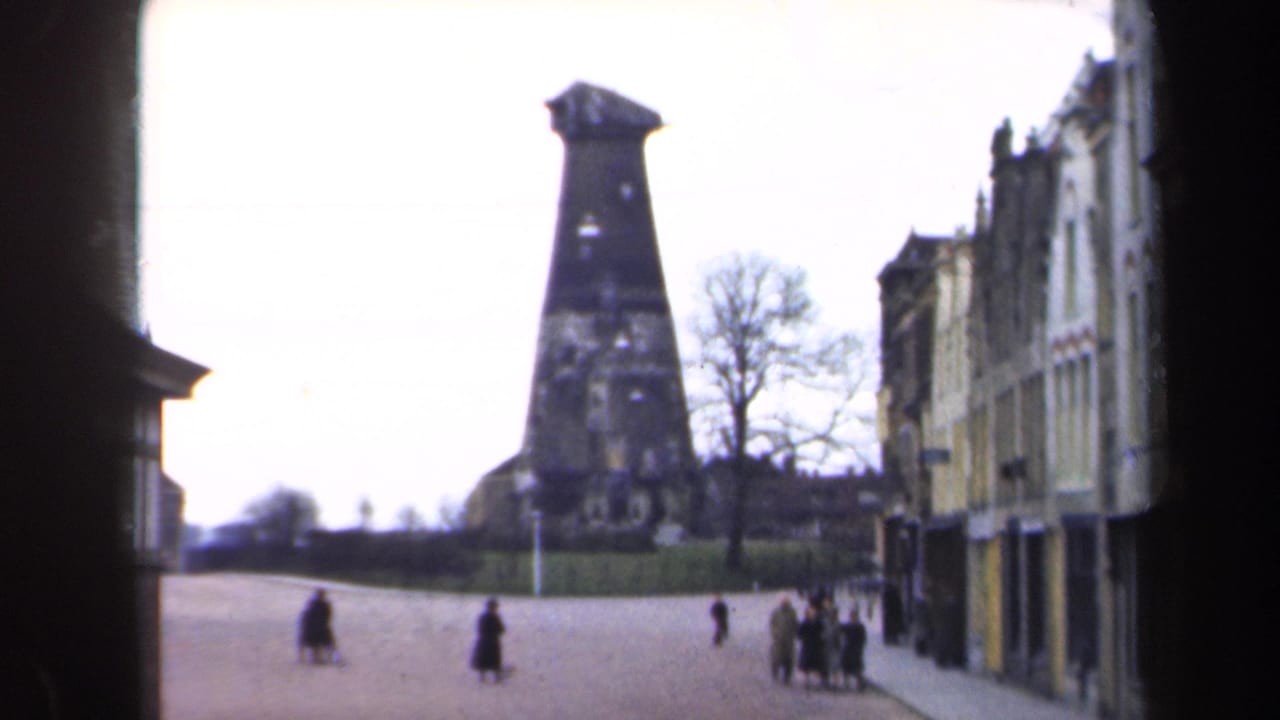 1946 Netherlands 8mm frame showing a quiet boulevard, pedestrians, building facades, bare trees, and a dark tower-like landmark.