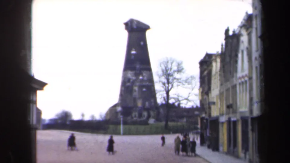 1946 Netherlands 8mm frame showing a quiet boulevard, pedestrians, building facades, bare trees, and a dark tower-like landmark.