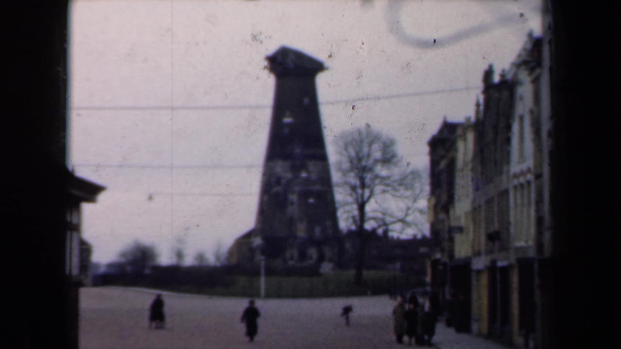 Pedestrians cross an open street beneath a tall dark landmark, with buildings lining the right side.