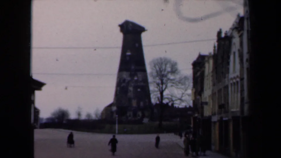 A blurred 1946 Netherlands 8mm frame holds on pedestrians in a broad street near a dark tower-like landmark and traditional facades.