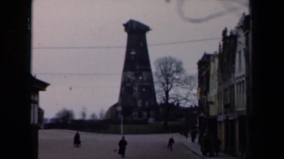 1946 Netherlands 8mm frame showing pedestrians on a wide boulevard, ornate facades at right, bare trees, and a dark tower-like landmark.
