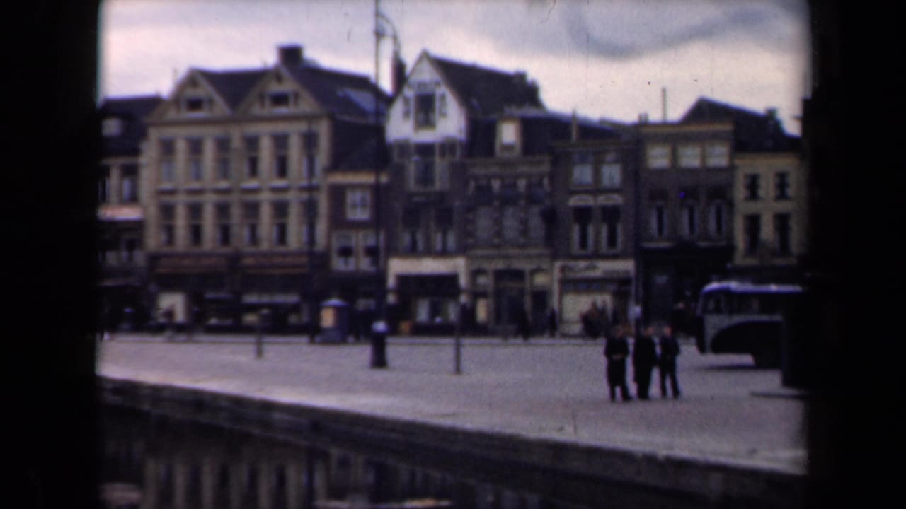 1946 Netherlands 8mm frame showing a broad riverfront quay, gabled urban buildings, small pedestrian groups, and a bus at the right edge.