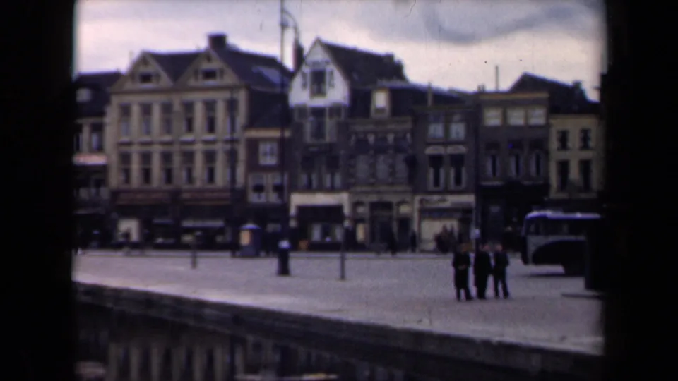 1946 Netherlands 8mm frame showing a broad riverfront quay, gabled urban buildings, small pedestrian groups, and a bus at the right edge.