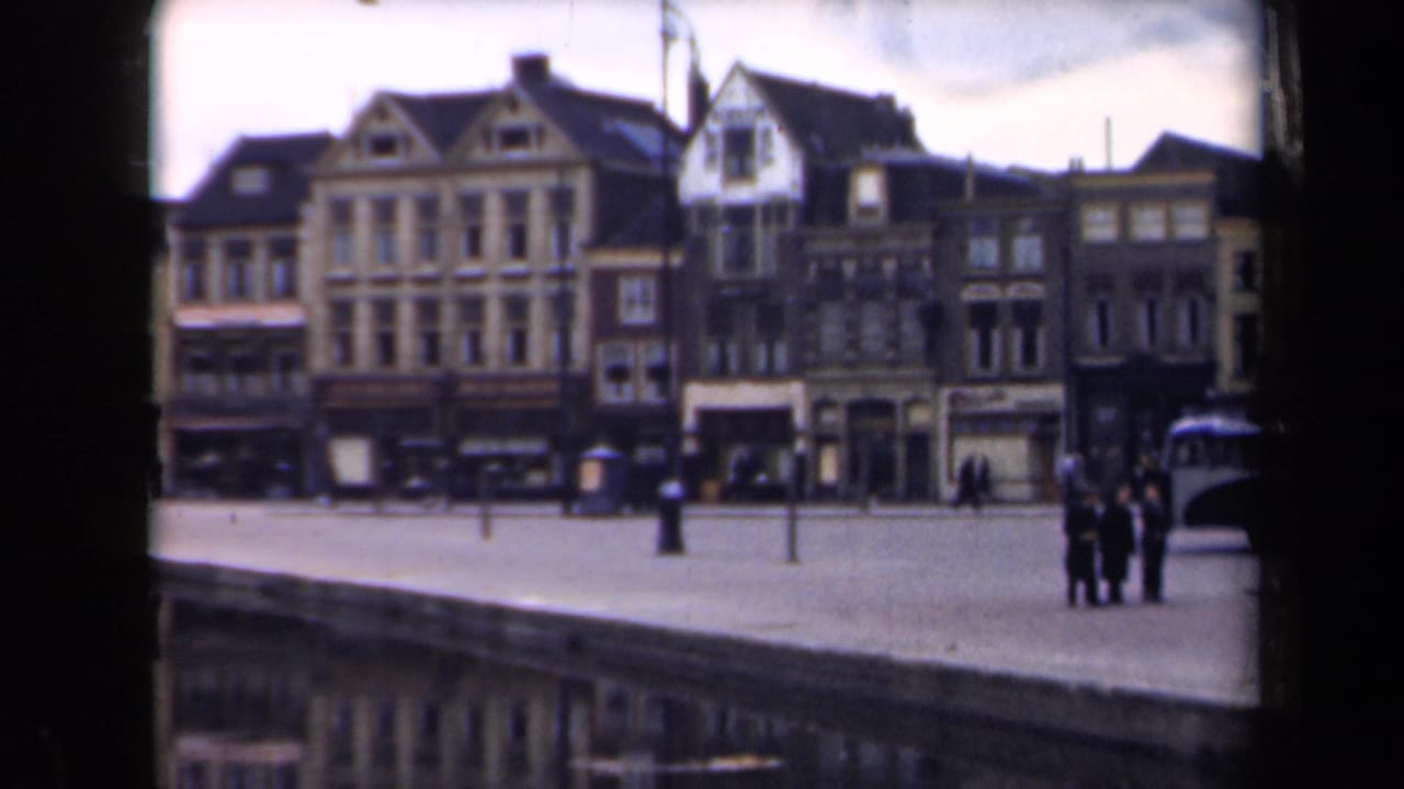 Waterfront street with gabled buildings, open quay, pedestrians, posts, and partial bus at right.