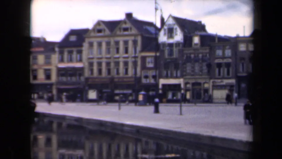 1946 Netherlands 8mm frame centered on gabled waterfront buildings, shopfronts, a broad quay, and distant pedestrians near the river.