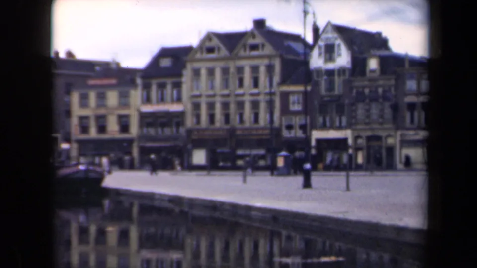 1946 Netherlands riverfront scene with water in the foreground, gabled buildings behind the quay, pedestrians, posts, and a partial boat-like form at left.