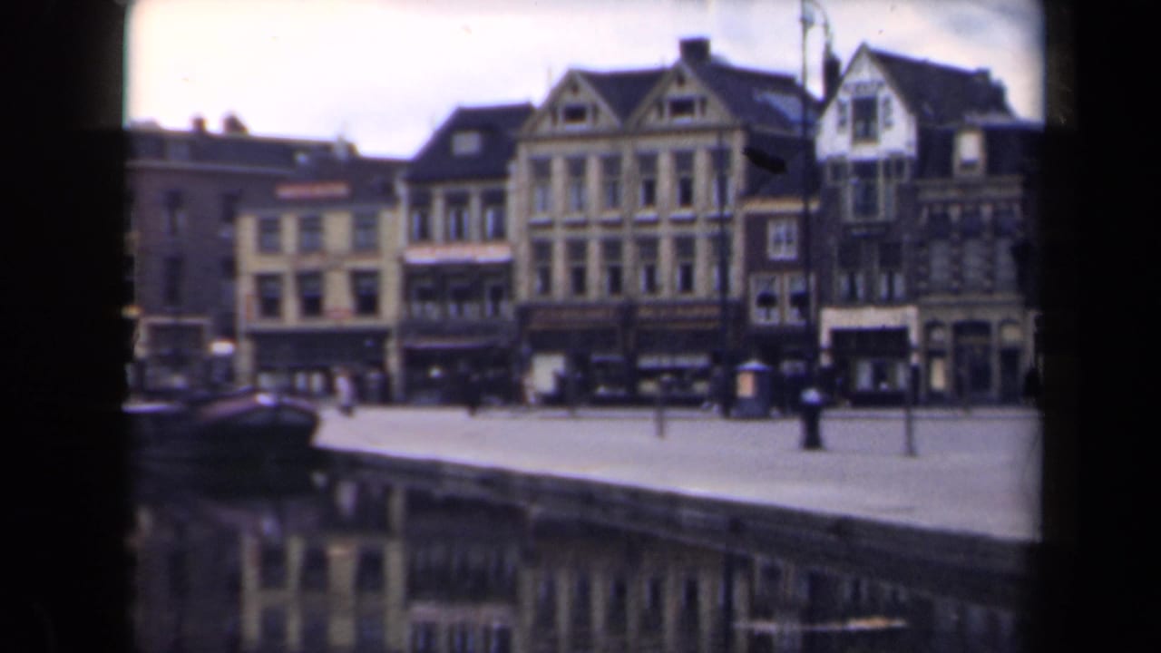 1946 Netherlands home-movie frame showing gabled canal-side buildings, a quay, pedestrians, and a dark moored boat.