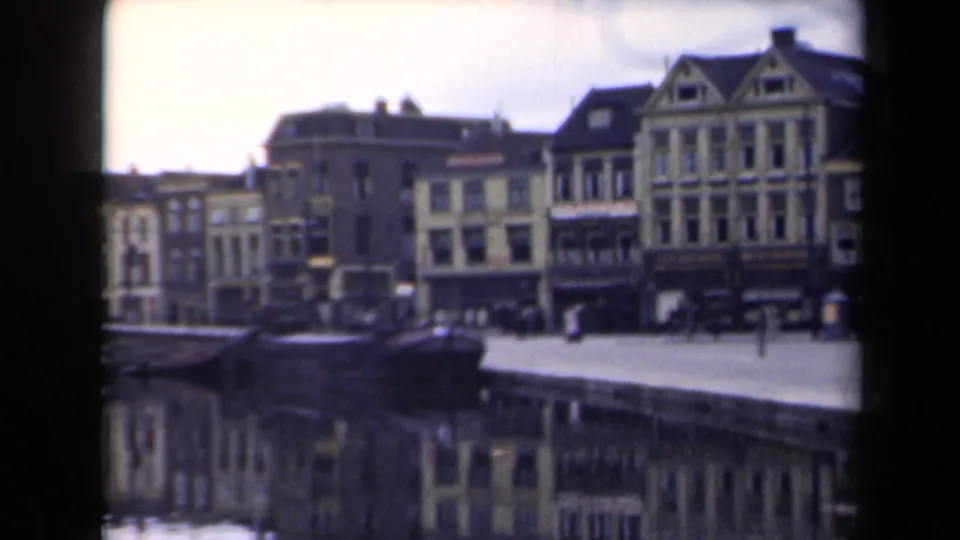 1946 Netherlands frame showing a canal edge, dark boat, pedestrians, and historic multi-story buildings across the water.