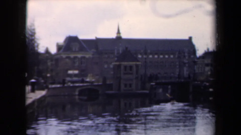 Soft 8mm frame of a 1946 Netherlands canal, with a large brick building, spire, stone quay, and water reflections.
