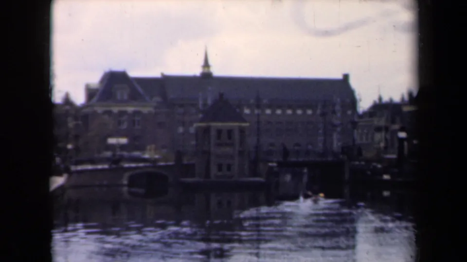 1946 Netherlands canal-side view with pedestrians, a bridge opening, rippled water, and a large brick building framed by 8mm gate edges.